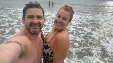 Anna Thomas A selfie of Dan (left) and his partner Anna (right) standing in the sea. Dan has short, dark hair and and Anna's red hair is tied back. They are both smiling. There are two people in the background also in the sea.