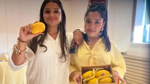 Aisha Iqbal/BBC Two young girls standing next to each other. One holds a single ripe mango, the other holds a box of four. Both wear yellow clothes.