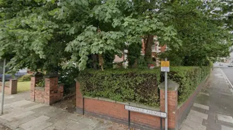 A brick wall covered in shrubs on a corner of a road. A street sign, reading Granville Avenue, sits in front. Trees cover the property behind the wall.