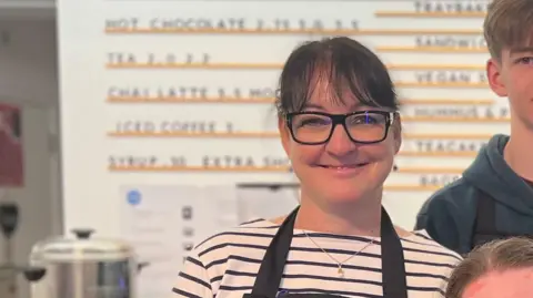 A woman with glasses smiling, she is wearing a stripy top with an apron over the top.  