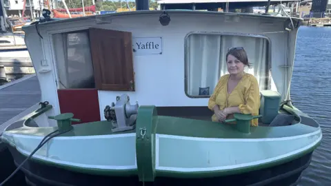 George King/BBC A woman wearing a yellow top standing on her green barge named Yaffle, which is docked in a marina.