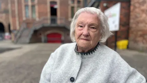 A lady with grey hair and a grey cardigan sits in front of some old buildings at the colliery site.