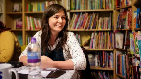 RNIB RNIB social media lead Becky Brynolf sitting in front of some bookshelves. She's smiling, wearing a white patterned blouse and pinafore dress.
