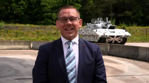A man in glasses, a navy suit and a blue striped tie stands on a concrete surface. In the distance behind his shoulder is an armoured vehicle.