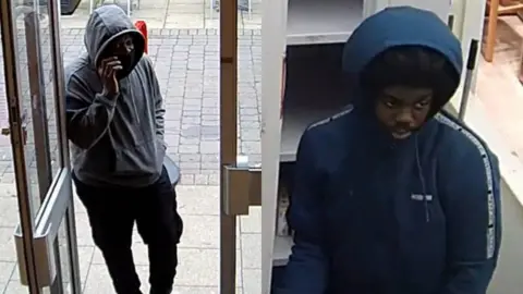 Two youths caught on CCTV committing a robbery in a mobile phone shop. One is wearing a blue hooded coat and the other a grey hoody and black trousers, both with their hoods pulled up.