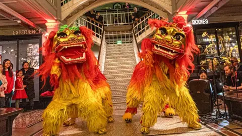 Manchester Museum Two gold and red Chinese dragons performing as part of Chinese New Year celebrations at the bottom of a staircase in Manchester Museum. Children watch to the left.