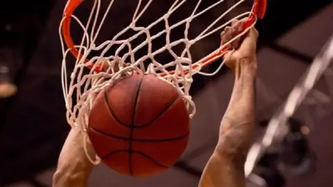 Getty Images A person holds on to a basketball net as a orange and black ball is pushed through, with the background appearing to be of a sports arena.