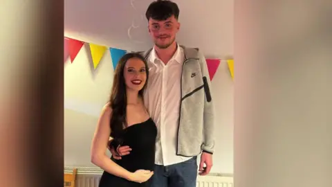 A young man and a young woman standing side-by-side with colourful bunting behind them. He is wearing a white, button-up shirt with a grey jumper over the top, and she is wearing a black dress and is holding her baby bump.