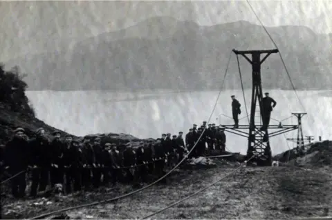 Scottish Water Archive photo of a workers lined up alongside a pulley system for taking materials from the Inversnaid area of Loch Lomond to Loch Arklet, where a dam was built as part of the Katrine Aqueduct project