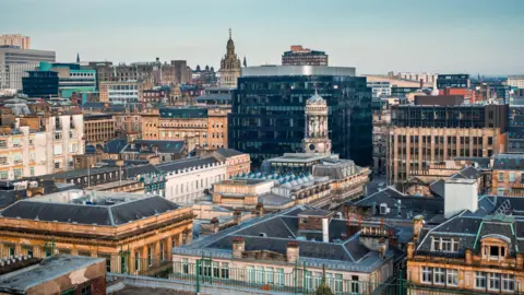 Getty Images Panoramic view of a dense city centre skyline featuring a mix of historic sandstone buildings and modern glass office blocks under a pale blue sky. Rooftops fill the foreground, while prominent church towers and civic buildings rise above the urban landscape in the distance. The scene is brightly lit by daylight, highlighting the contrast between traditional and contemporary architecture.