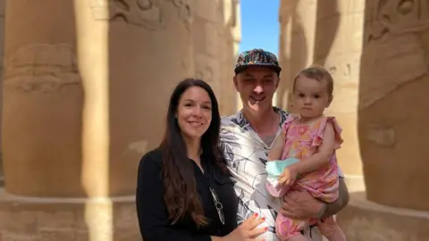 Family Handout Paul and Ally standing between sandstone columns. Paul is holding their daughter who is wearing a pink dress. 