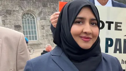 Nosheen Khalid A woman smiling in front of a building in a city square. She is wearing a black hijab covering her hair and a navy coat. Behind her the shoulders of others standing in her group can just be seen.