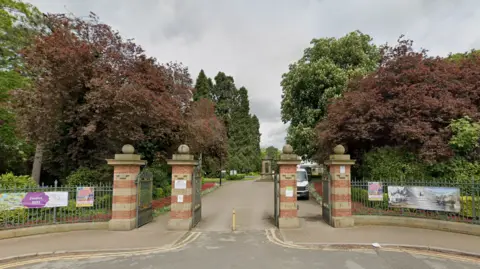 The entrance to South Park has four red brick columns with dark green metal gates between them. Lush green and brown trees can be seen in the park within.