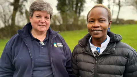 BBC Two women stand side by side in a green field. An Irish woman with short grey hair looks at the camera. She is wearing a navy fleece and navy T-shirt. She is standing beside a woman from Rwanda who has short dark hair and long earrings. She is wearing a white top with the black puffer jacket over it.
