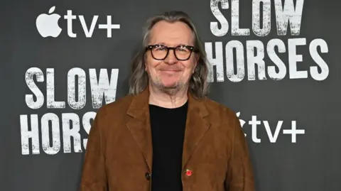 Getty Images Gary Oldman is wearing a brown jacket and black jumper underneath. He has long mostly grey hair and is unshaven resembling his Slow Horses character Jackson Lamb. He is at an event promoting Slow Horses and standing next to the show's logo.