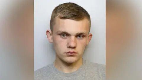 A police custody photo of Bert Baker. He has short brown hair and is wearing a grey t-shirt.