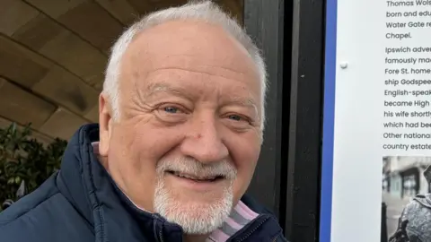 A man with grey hair is smiling at the camera. He is wearing a navy gilet jacket with a navy jumper underneath and a white top. He is standing in front of a town information board detailing its history and points of interest.