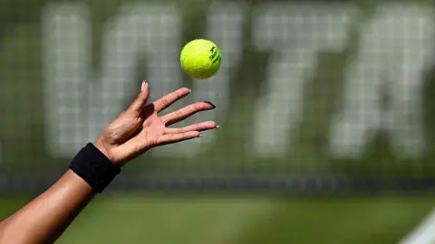 A female tennis player with long fingernails (only the hand shown) tosses a tennis ball in front of a net featuring WTA