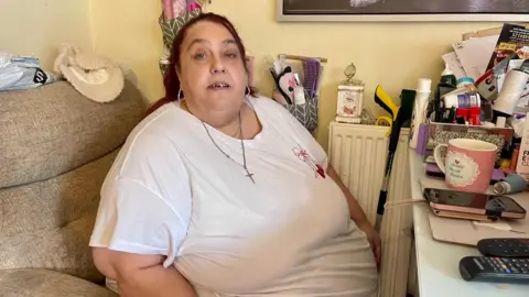 A woman in a t-shirt with long dyed red hair is sitting in an armchair in front of a desk. He has large earrings and a chain with a crucifix. The table has a mug, remote control, phone, tablets, inhaler and paperwork on it. She has a metre-long grabber beside her resting on a radiator. There is a carriage clock and a small basket of brushes and make up by her side.