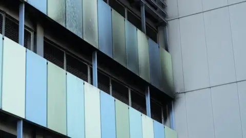Shaun Whitmore/BBC Part of the outside of a car park shows black charring on green and blue panels and a wall.