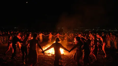 ANASTASIA FREEMAN Performers dancing around a fire while holding hands.