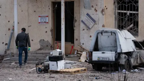 A man walks towards a building damaged by a Russian strike, past a car wreckage and office equipment piled amid debris in a yard on 24 March 2026 in Poltava, Ukraine