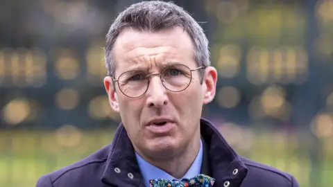 Andrew Muir is standing outside on front of large black gates. He wears glasses and is wearing a navy coat with a blue shirt and a bow tie.