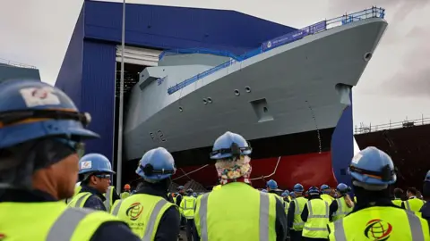 The Royal Navy frigate, HMS Venturer, being rolled out of the build hall at Babcock Rosyth. Dozens of workers in hi-vis jackets and blue helmets are watching with their backs to the camera 