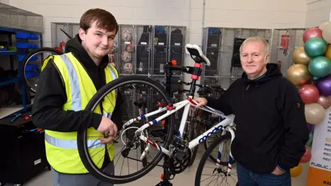 Two men standing in a bicycle repair shop. A white road bike is being held on a repair stand.