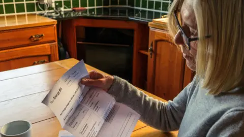 Getty Images Stock image of a woman reading an energy bill. The woman has blonde hair and glasses and is wearing a grey jumper. She is sat at a kitchen table with a cup of coffee. 