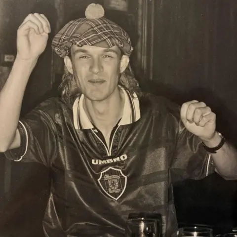 Colin Paterson Black and white image of a man in tartan tammy with his hands raised in celebration. He is wearing a blue Umbro Scotland home top.