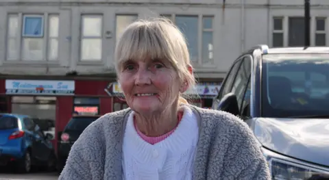 Jose Wilmin sits on a bench on the promenade at New Brighton. She is in her late 60s and is wearing a white cardigan and fluffy grey fleece. 