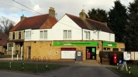 A picture of the Horspath post office. A green Londis banner sits above the window and a red postbox stands outside the door