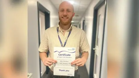 Bradford Teaching Hospitals NHS Foundation Trust Alex Overton is smiling as he stands in a corridor holding his certificate. It says 'certificate of stem cell donation' and includes the words 'thank you'.