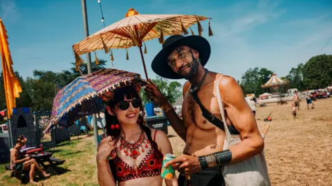 Shinding Festival A man and a woman pose for the camera at the Shindig Festival, each of them holding a parasol to keep off the sun. She is wearing a red patterned crop top, a large red necklace and sunglasses. He is wearing a black hat and no top.