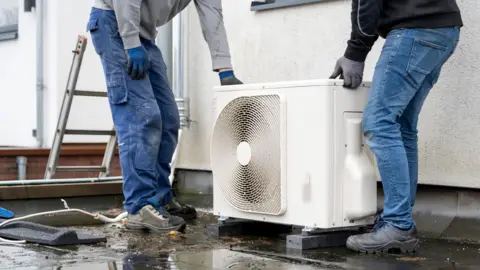 The shot shows a white heat pump being installed on a wet flat roof by two men in jeans and boots. Just the bottom half of their bodies are visible as they fit the pump into place. A ladder is visible just to the left of the picture.
