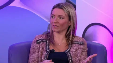 Francesca O'Brien is sat in a chair in front of a microphone wearing a red cardigan and a blue top.