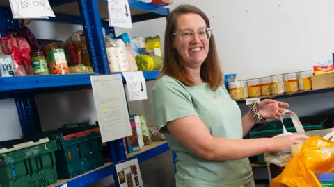 Cumbria Community Foundation Steph Humes has medium-length brown hair and clear framed glasses. She is wearing a teal-coloured t-shirt and is packing food into a plastic bag at a foodbank. 