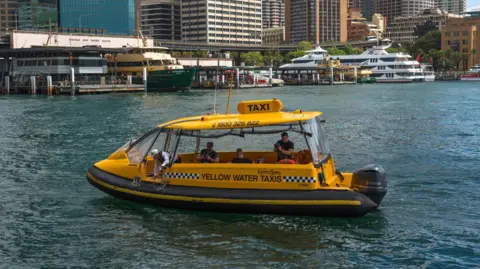 Image shows a yellow water taxi with Sydney waterside in the background. There are three people on board and a driver.