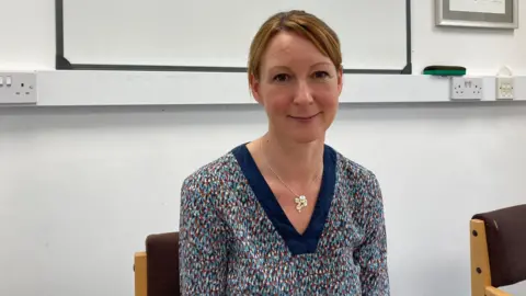 Matthews sat on a chair in a room with white walls. She is wearing a patterned top of multiple colours and is wearing a silver necklace. She has long ginger hair, which is tied back. 