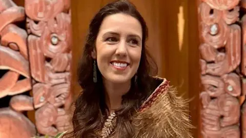 J Ancell Laura wears a feathered cloak. She has long brown hair and is smiling. She is pictured in front of a decorative wall featuring Māori carvings.