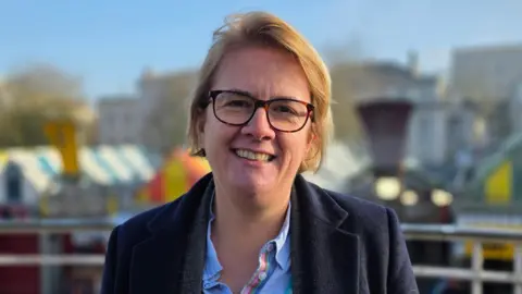 Paul Moseley/BBC Carli Harper standing by the war memorial on the top of Norwich Markets in the city. Behind her is colourful striped canopies of market stalls. Carli Harper is looking directly at the camera and smiling and is wearing a blue shirt with a coat over the top.