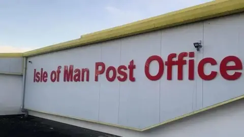 The roof of the post office headquarters in Douglas, which shows the top of the white building that has Isle of Man Post Office written on it in red lettering below the yellow roof.