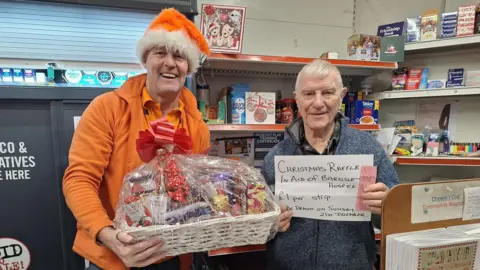 Gordon Bird A man in an orange fleece and Santa hat stands behind a counter smiling and holding a large hamper. An older man in a dark grey cardigan stands next to him