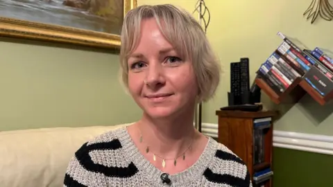 Mina has short blonde hair. She is sitting in her living room on an armchair and smiling at the camera.