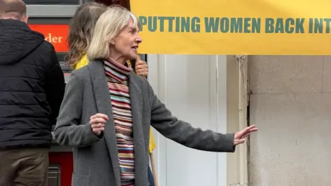 Chichester Women's History Group A woman stands on a pavement and gestures while speaking, with part of a yellow banner visible behind.