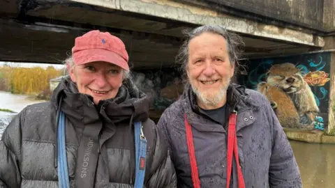 A man and a woman are smiling a the camera, whilst standing next to a river in the rain. The woman is on the left and is wearing a black coat and a pink baseball cap. The man has a grey beard and long dark grey hair, tied back. He is wearing a dark blue coat. In the background is a painted mural of otters, on the wall under a bridge.