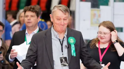 Athena Pictures/BBC Andrew Slaughter is smiling with two other people stood next to him. He is wearing a Green rosette
