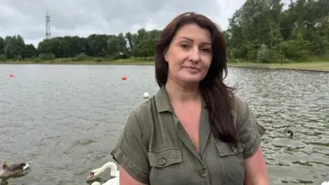 Blyth Wildlife Rescue Centre Sara Perry, with long brown hair, wearing a green khaki dress, looks at the camera as she stands in front of a large pond. Swans and ducks are on the pond behind her. It is a cloudy day.