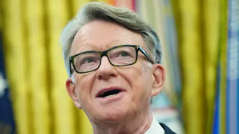 Lord Mandelson in the White House Oval office. he is mid-speech and is wearing  black-rimmed glasses. It is a close shot of just his face. In the background, are gold curtains.
